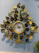 A decorative wall clock shaped like a tree, with a gold-toned metallic trunk and branches made from mirrored leaves. The clock face is white with black numerals and hands, mounted against a light blue-gray wall.