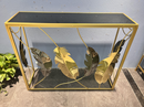 A rectangular console table with a gold metal frame, black mirrored top and base, and large decorative feather motifs in gold and silver tones, shown against a light blue wall.