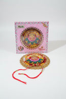 A decorative gold-plated rakhi with colorful embroidered details and a red hanging cord, displayed on a wooden surface below its pink retail box.