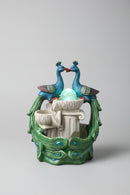 A decorative peacock-themed fountain sculpture with two blue peacocks facing each other above a white pedestal, surrounded by green peacock-feather-like forms with blue eye details.