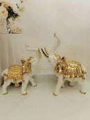 Two white elephant figurines decorated with ornate gold saddles and trinkets, posed facing each other on a light surface against a white background.